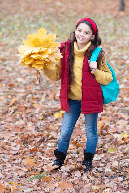 Schoolgirl holding autumn leaves outdoors Happy girl walking in autumn park after school