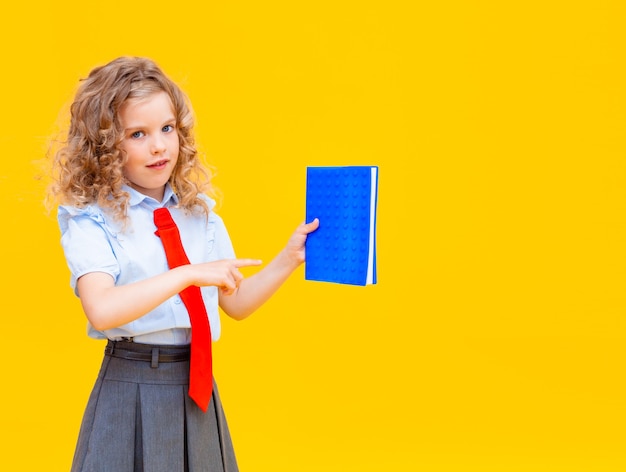 Schoolgirl holds an open blue notepad. Pupil with long braids isolated on a yellow background.
