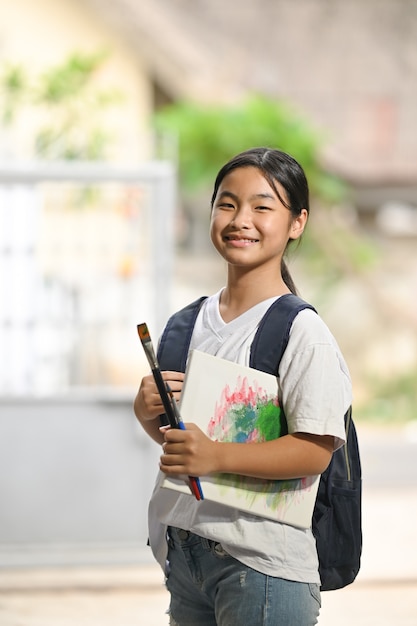 A schoolgirl is holding painting equipment and carrying a school bag while standing and waiting for a school bus.