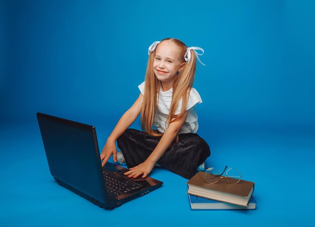 A schoolgirl is sitting on a blue background and looking at a laptop a little girl is studying online the child is typing on a laptop isolated background