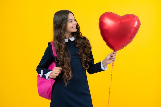 Schoolgirl on the last day of school with balloons i love\
school school girl with school bag hold heart balloon isolated on\
yellow background