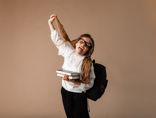 A schoolgirl looks at the camera, holding a book, and showing her tongue, straightens her glasses. Education concept.