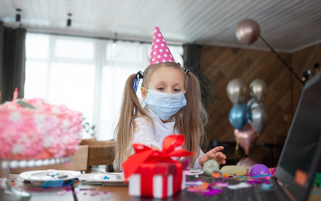 医療用マスクとお祝いの帽子をかぶった女子高生がテーブルの近くに立っています。女の子は贈り物を見ます。