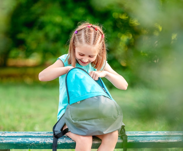 Schoolgirl peeking into open backpack on bench in park