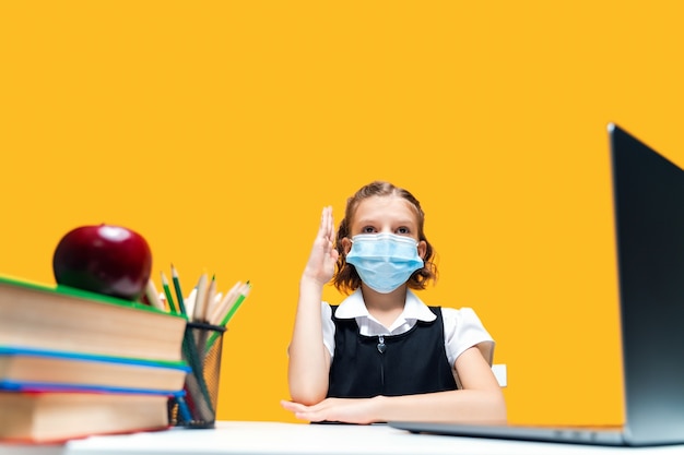 Schoolgirl raising hand and sitting at laptop wearing mask yellow background distance learning