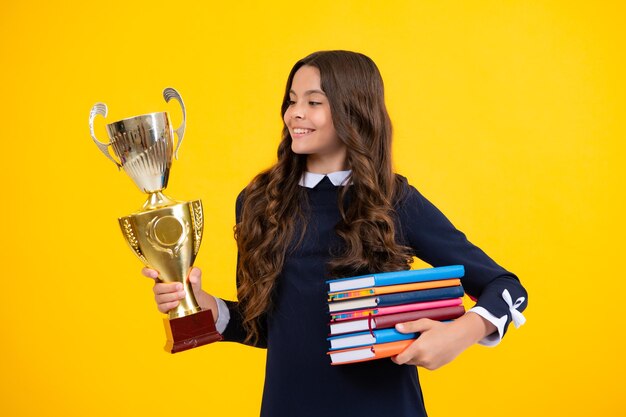 Schoolgirl in school uniform celebrating victory with trophy teen holding winning award prize on yellow background child hold books with gold trophy or winning cup education graduation victory