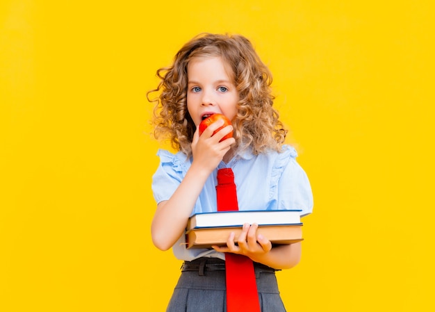 Schoolgirl in a school uniform holding a stack of books and a red apple on a yellow background. Concept of learning and school.