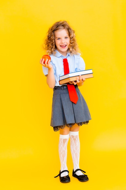 Schoolgirl in a school uniform holding a stack of books and a red apple on a yellow background. Concept of learning and school.