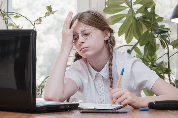 Schoolgirl self isolation using laptop for his homework. Gaming, education and school concept - sad girl with laptop and at home. Girl having hard time doing homework online, sitting depressed indoors