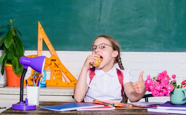 Schoolgirl sit desk chalkboard background Kid student in school Girl small child eating apple snack School break Relax between lessons Vitamin charge School life concept Healthy lifestyle