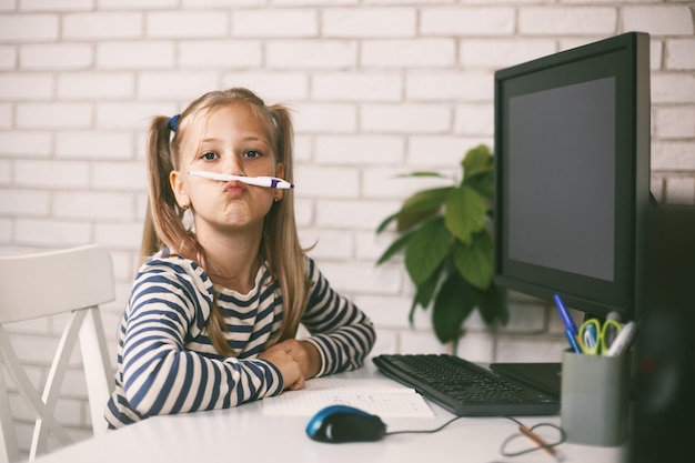 La studentessa si siede a casa al tavolo davanti al computer, gioca con una matita, gioca e ride.