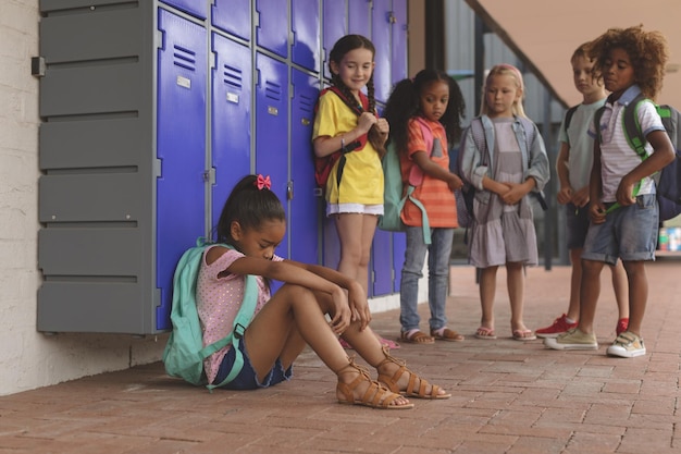 Schoolgirl sitting alone in school corridor while others school kids looking at her