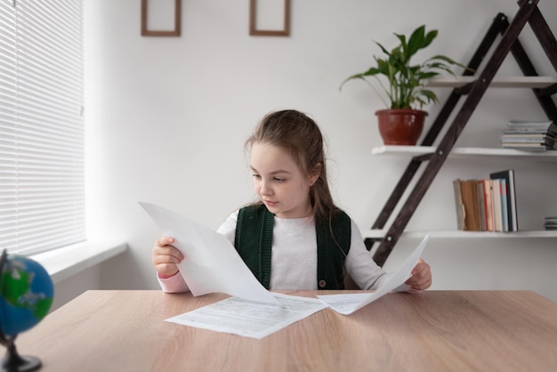 Schoolgirl student girl sits at home and learns online distance\
pupil is holding sheets of paper with homework in his hands and\
thinking intently over the answers to the teacher video call