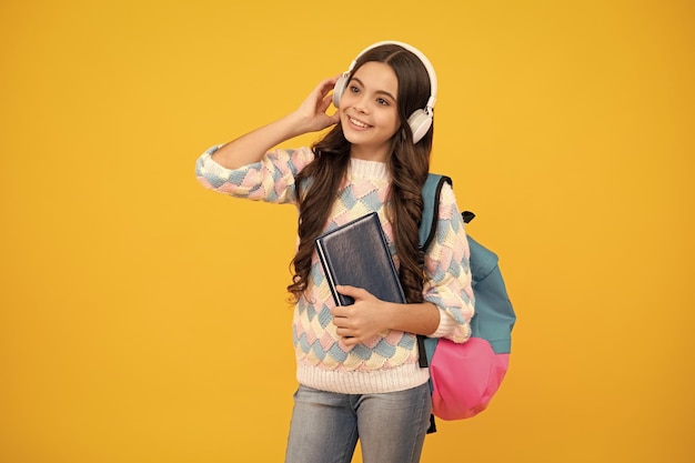 ヘッドフォンで女子高生の10代の学生の女の子は、黄色の孤立したスタジオの背景に本を保持します学校