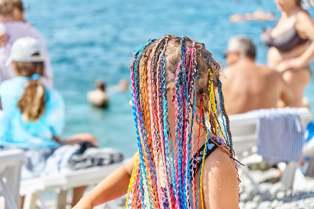 Schoolgirl tourist with fake colourful dreadlocks on crowded sea beach