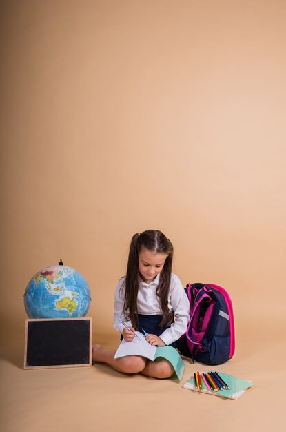 A schoolgirl in a uniform is sitting with school supplies writing in a notebook on a beige background with a place for the text