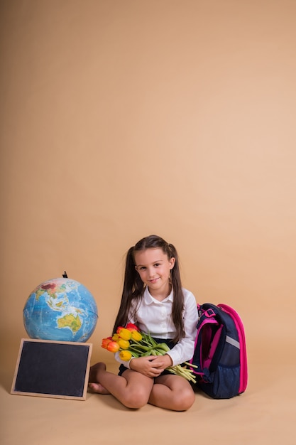 A schoolgirl in a uniform sits with school supplies on a beige background with a place for text