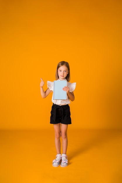 Schoolgirl in a uniform stands and holds a blue notebook on a yellow background with a copy of the space. At full height