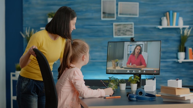 Schoolgirl using video call for online lesson with teacher and mother at home. Parent giving assistance to young child while attending school courses with video conference on computer.