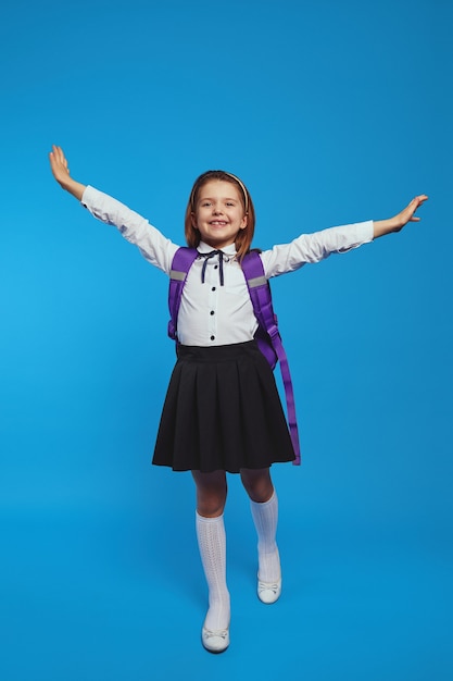 Schoolgirl wearing uniform and backpack raises hands up against blue wall