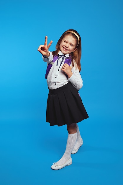 Schoolgirl wearing uniform and backpack smiling while showing peace gesture