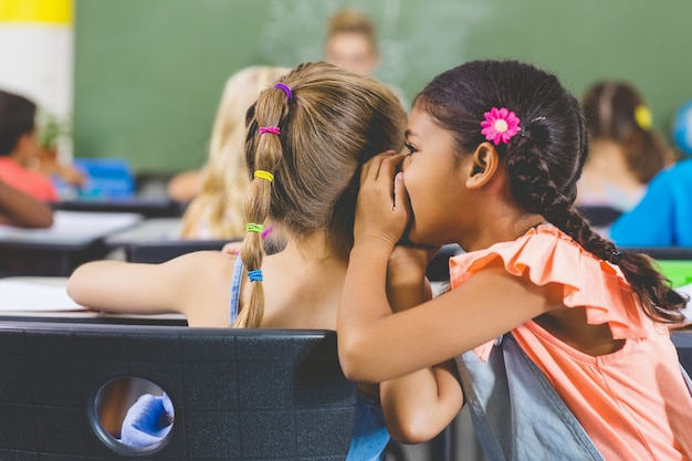 Schoolgirl whispering into her friend s ear in classroom