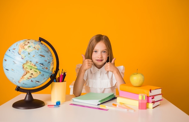 Schoolgirl in a white blouse is sitting at a table with school supplies and a globe and shows the class on a yellow background with a copy of the space