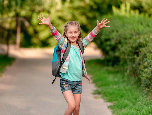 Schoolgirl with backpack going to school with hands up on park background