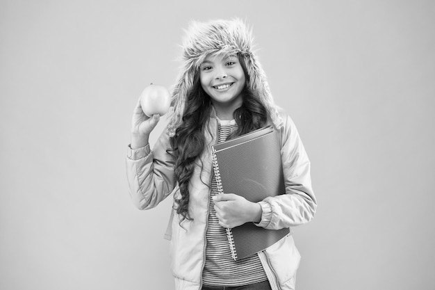 schoolgirl with books after lesson school break small happy girl earflap hat eat apple winter holiday vacation teen kid pink background pupil daily life Modern education Student of the month