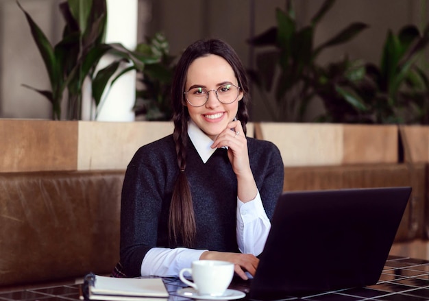 Schoolgirl with braids wearing eyewear sitting with coffee and laptop in the wooden cafe