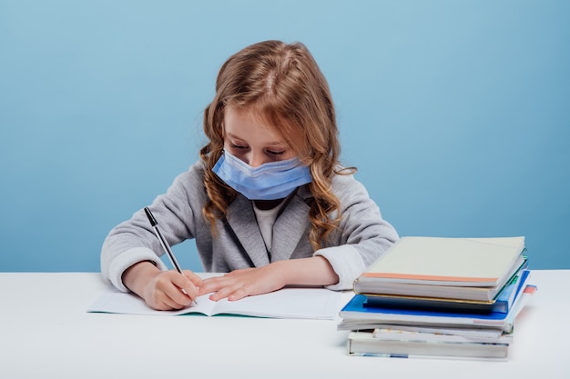 Schoolgirl with medical mask writes in notebook sitting at the table isolated on blue background