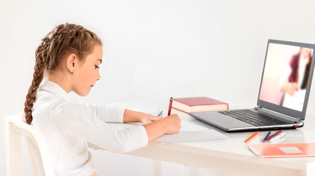 Schoolgirl writes in a notebook sitting at a table in front of a laptop