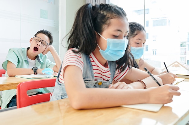 Schoolgirls in medical masks writing in copybooks when their classmate yawning