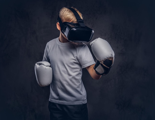 Schooljongen bokser met blond haar gekleed in een wit t-shirt met een visuele realiteitsbril en bokshandschoenen, trainen in een studio. Geïsoleerd op een donkere gestructureerde achtergrond.