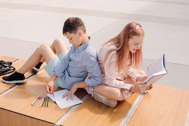 Schooljongen en schoolmeisje zitten rug aan rug op een bankje op het schoolplein bij het schoolgebouw. studeer met boeken en schriften