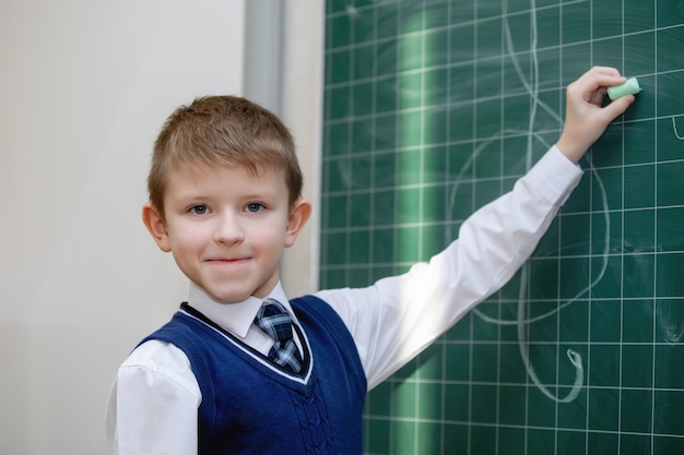 Schooljongen in uniform schrijft met krijt op een schoolbestuur. school basisonderwijs. selectieve aandacht.