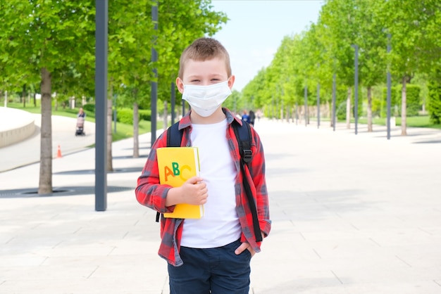 Schooljongen met een beschermend masker tegen het virus, met een leerboek in zijn handen, staat op straat op weg naar school. Terug naar school