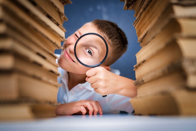 Foto schooljongen zit tussen twee stapels boeken camera kijken door het vergrootglas.