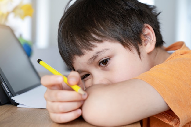 Schoolkind in zelfisolatie met behulp van tablet voor zijn huiswerk, Bored Child verdrietig gezicht liggend hoofd naar beneden te kijken diep in gedachten,