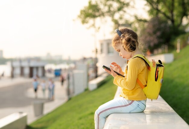 Schoolkind met rugzak in de natuur luistert muziek met mobiele telefoon en koptelefoon. Online technologiecommunicatie. Online onderwijs op de basisschool. Quarantaine is voorbij. Terug naar school.