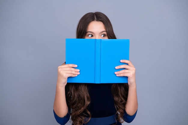 Schoolkind tiener student meisje houdt boek in de buurt van ogen op grijze geïsoleerde studio achtergrond School en onderwijs concept Terug naar school Gezicht verborgen met boek