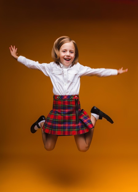 Schoolmeisje dat hoog springt en zich verheugt over de terugkeer van schoolkleding, casual geruite rok, geïsoleerde gele achtergrond