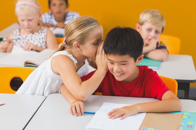 Foto schoolmeisje dat in het oor van haar vriend in klaslokaal fluistert