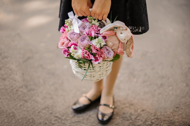 Schoolmeisje die een leuk rieten mandhoogtepunt van heldere roze bloemen houden die met een stuk speelgoed konijn worden verfraaid