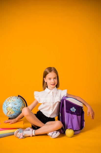 Schoolmeisje in uniform zit op de grond met schoolspullen op een gele achtergrond met een plek voor tekst