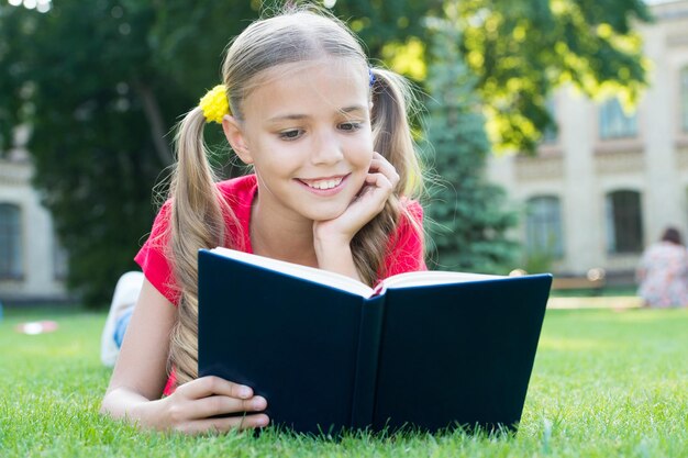 Schoolmeisje leest verhalen terwijl ze een groen gazon ontspant. Leuke leerling geniet van lezen. Schooltijd. Interessante verhalen voor kinderen. Studeer met plezier. Tijd voor mooie verhalen. Klein kind leesboek buitenshuis.