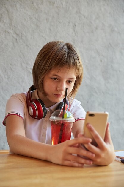 Schoolmeisje met koptelefoon en rode ijzige limonade gebruikt smartphone surfen op internet