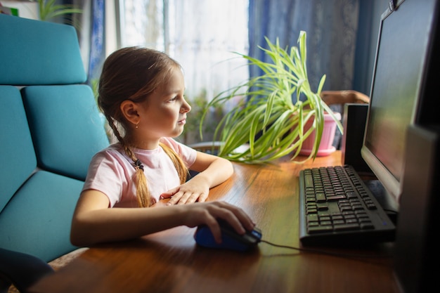 Schoolmeisje met staartjes zit thuis in een fauteuil op een desktopcomputer en bestuurt een computermuis