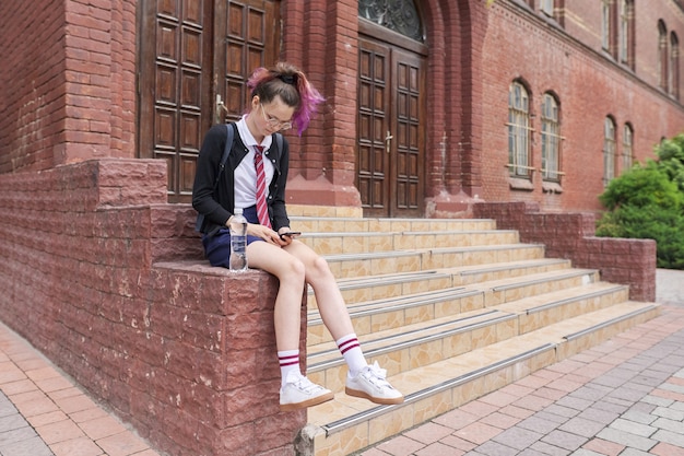 Schoolmeisje tiener in uniform met rugzak met smartphone. Meisje in de buurt van schoolgebouw, kopieer ruimte. Terug naar school, terug naar school, onderwijs, tieners concept