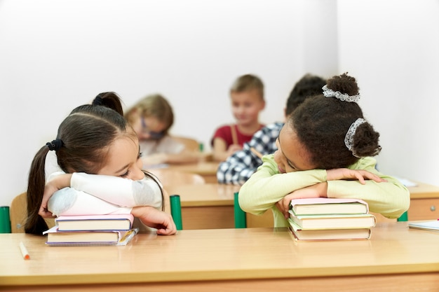 Schoolmeisjes die in klaslokaal bij bureau slapen, die op boeken leunen.